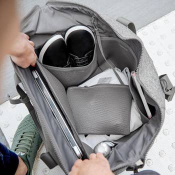 hands holding the duffle in grey open, showing how many pockets and space is inside, including a laptop sleeve, pocket for shoes, and smaller mesh pouch