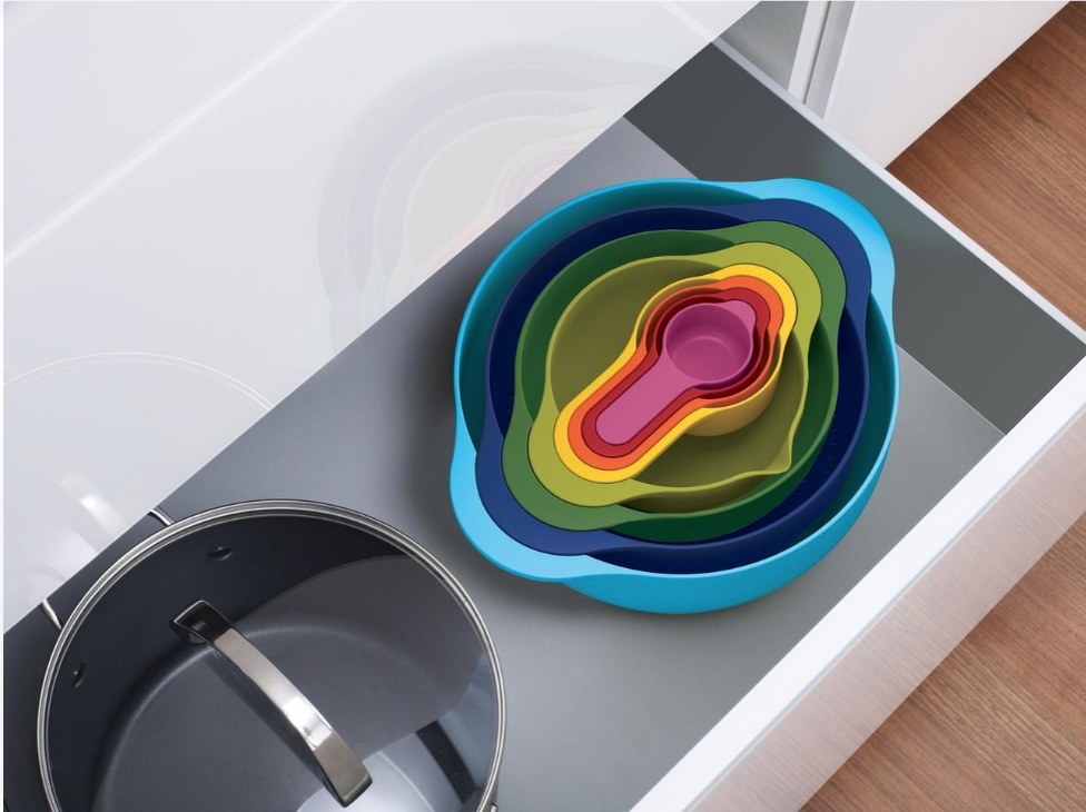 A colorful, stackable set of mixing bowls, measuring cups, and a colander displayed in a kitchen drawer