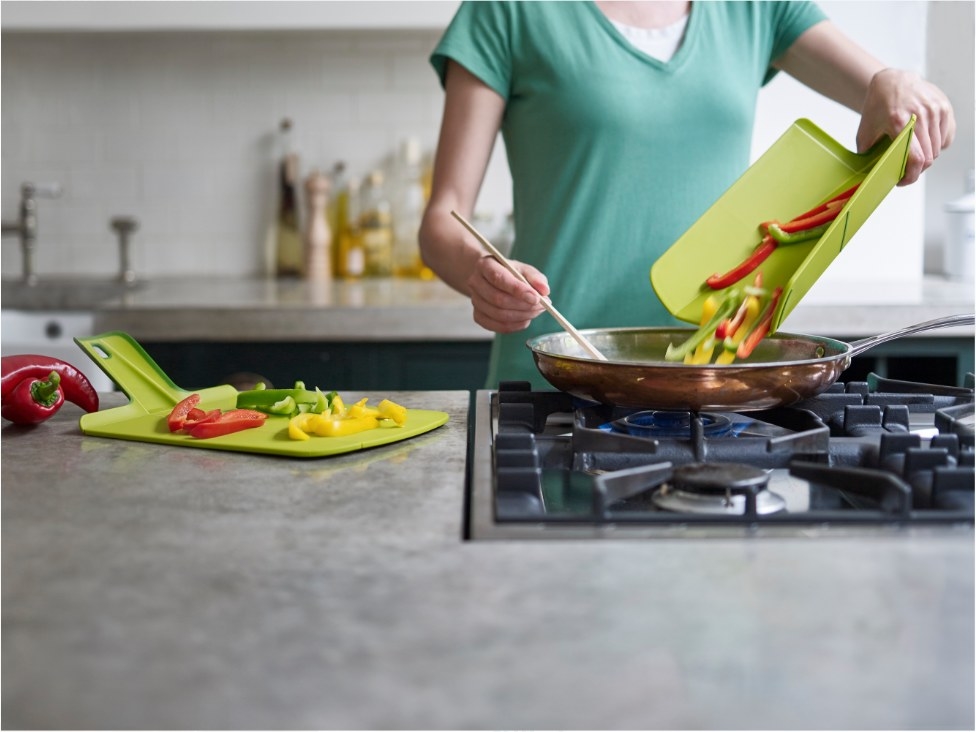 A model gripping the soft-grip handle of a green foldable chopping board that is being used to toss peppers into a pan