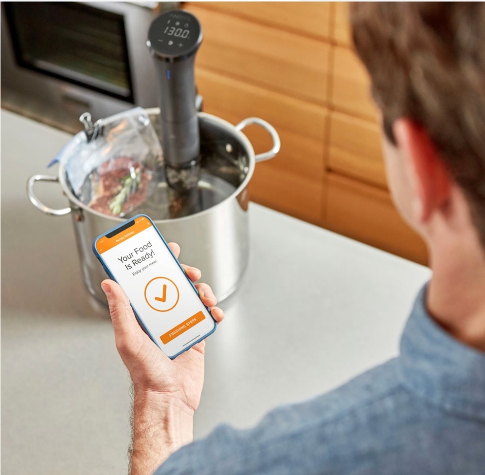 A model checking their foods progress on their phone while the bluetooth-controlled sous vide cooks the food in a pot on the counter