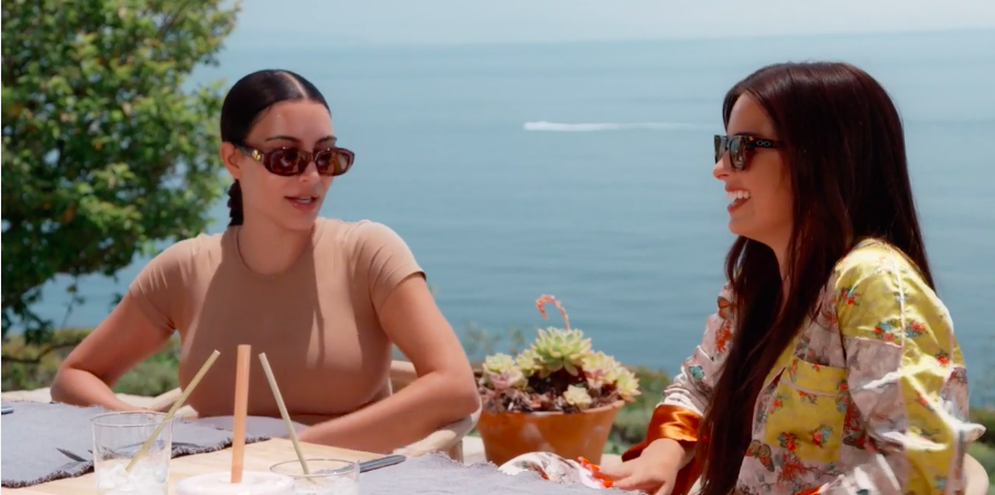 Kim and Addison wear sunglasses outdoors at a dining table by the ocean