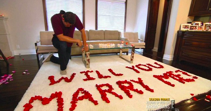 Kris kneels on a rug in front of rose petals spelling out the words &quot;will you marry me?&quot;