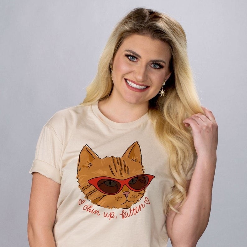 model wearing tan tee with orange cat face wearing red cat eye sunglasses above read script reading &quot;chin up, kitten&quot; with doodle hearts
