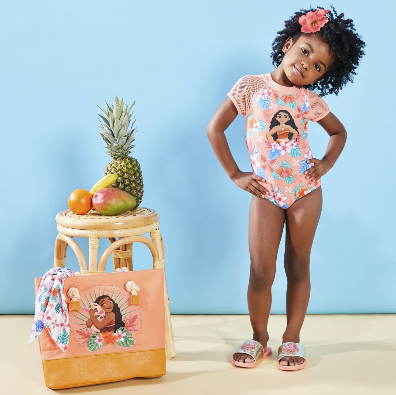 一个穿着泳衣的孩子站在莫阿纳大手提袋旁边，它的图案和毛巾一样，顶部是粉红色的帆布，底部是棕色的皮革