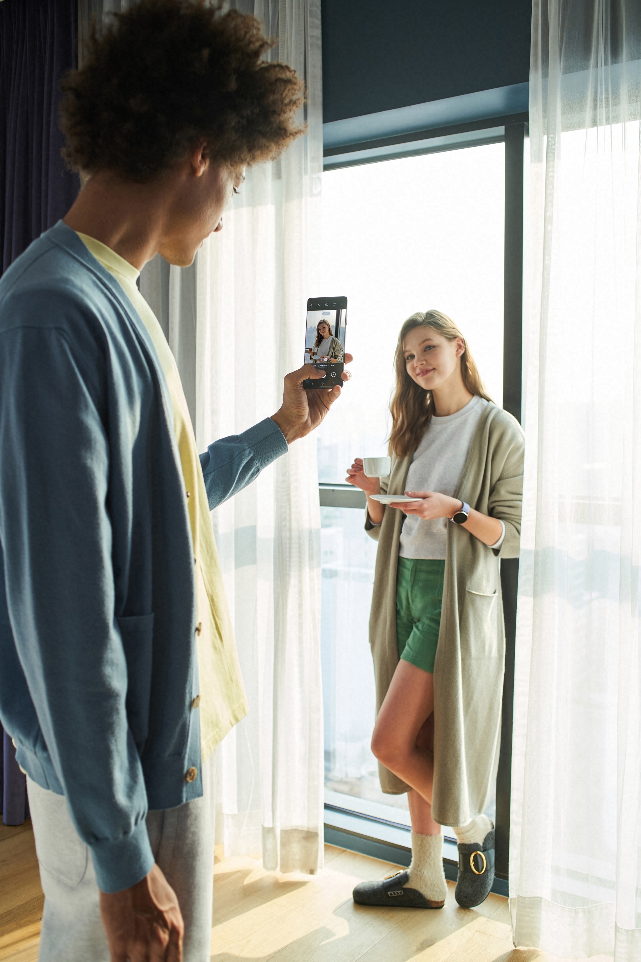A girl in a green robe poses with a cup of coffee beside a window as a boy takes a photo on his cellphone.