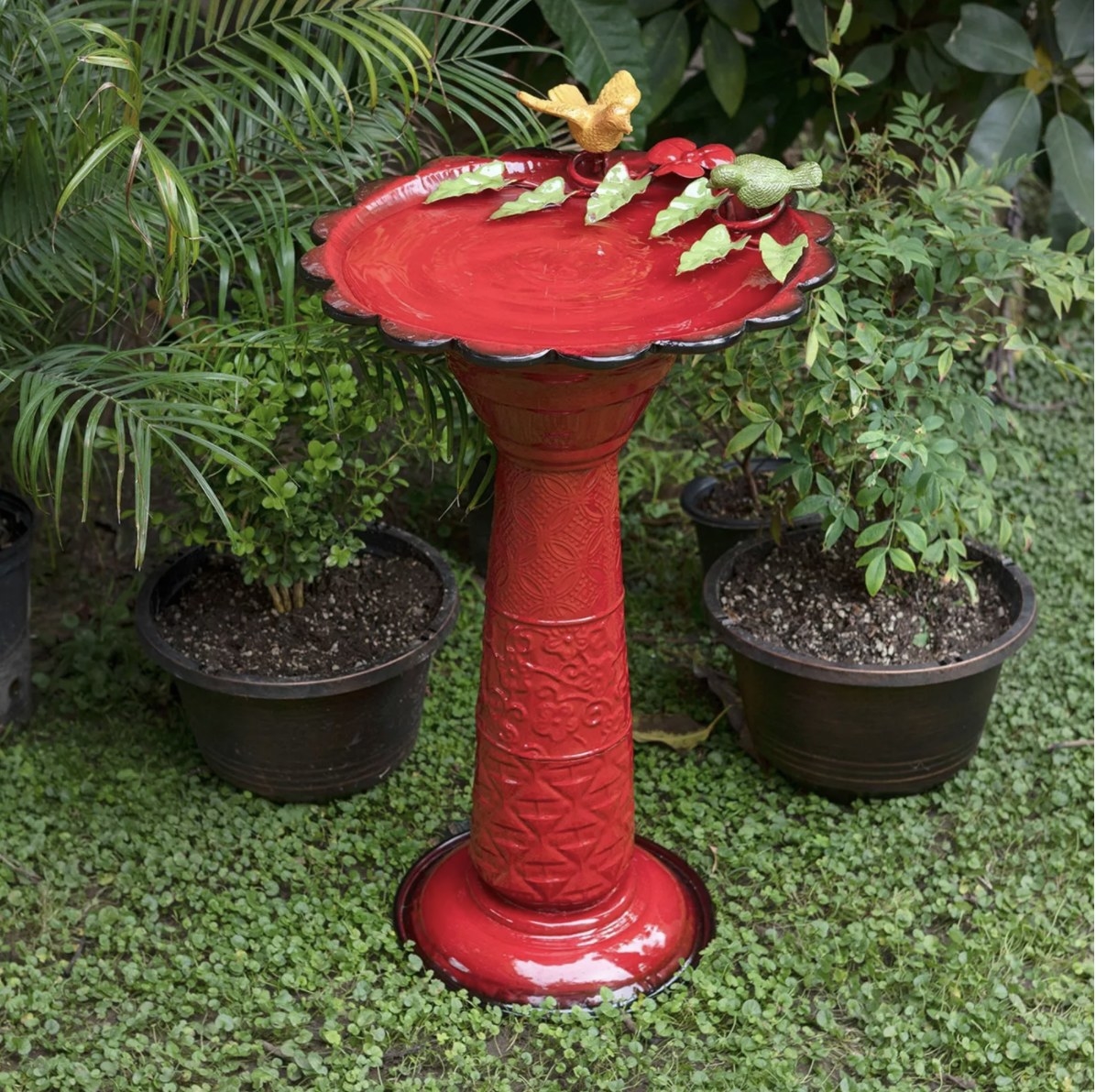 The bright red birdbath is outside surrounded by greenery and has two metal birds and some metal leaves on top 