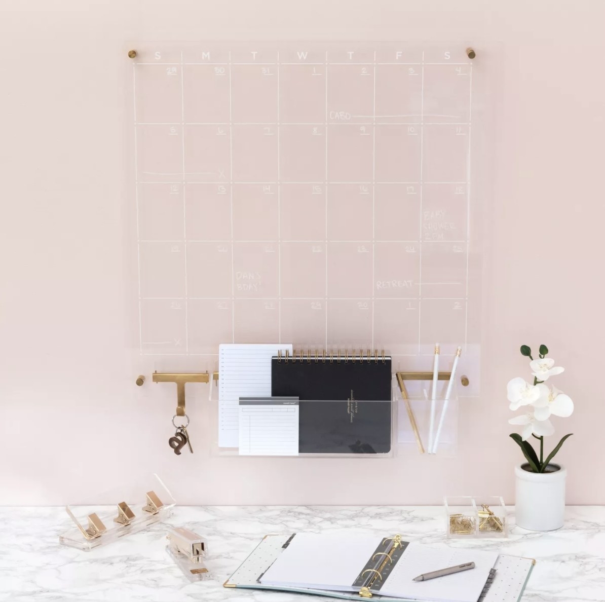 The acrylic calendar is hung on a light pink wall and surrounded by white and black decor and accessories