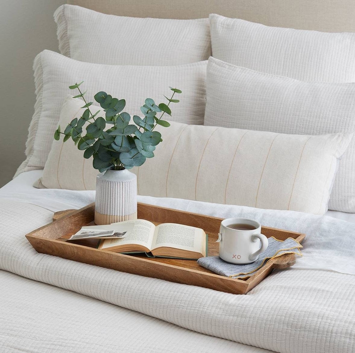 faux eucalyptus arrangement in a painted white pot sitting on a tray on a bed 