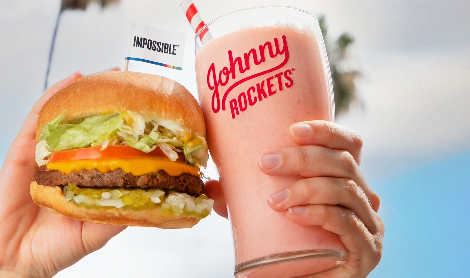 one hand holding a burger with an impossible flag on it and another hand holds a pink milkshake