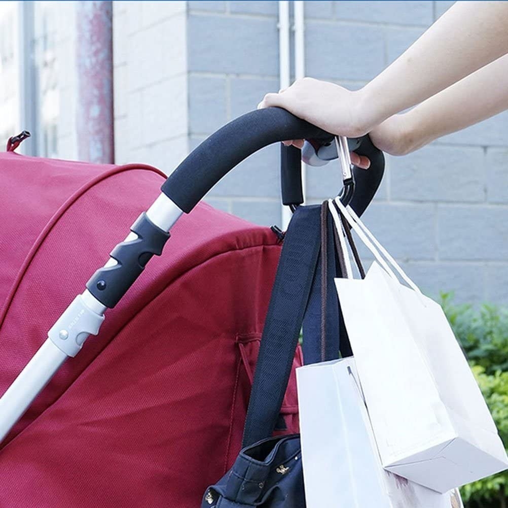 A stroller with a large carabiner hook attached to it A purse and multiple shopping bags are suspended from the hook