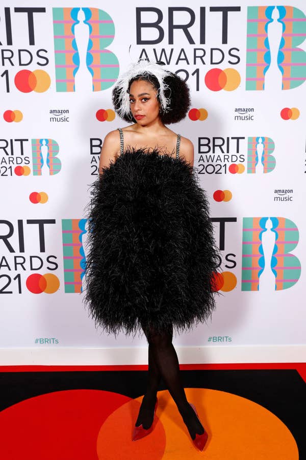 Celeste poses in a short feathered dress and stockings in the media room during The BRIT Awards 2021