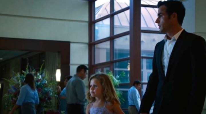 Kevin wears a suit and walks through a hotel lobby holding hands with a little girl
