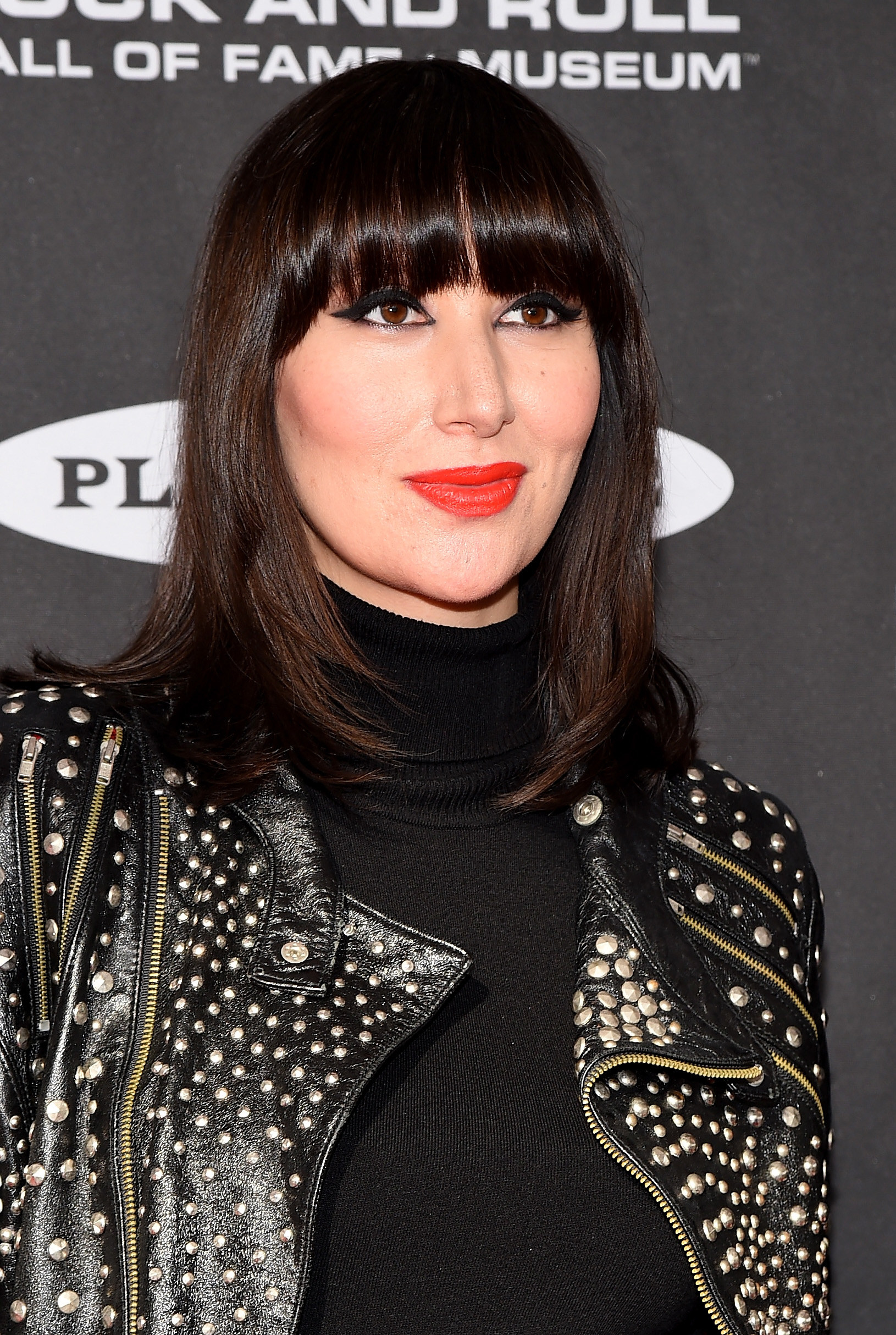 Karen O smiles with a bold red lipstick, black turtleneck, and leather jacket 