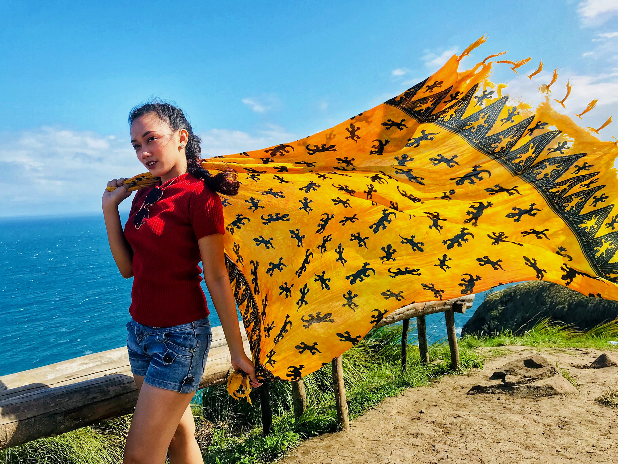 Woman in red shirt and jean shorts holding yellow scarf blowing behind her