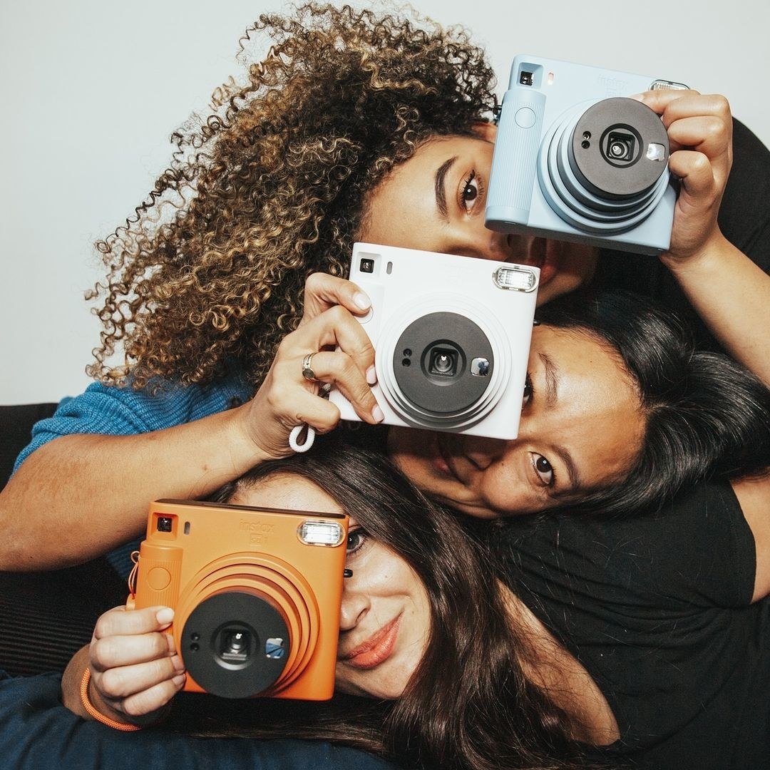 Three people leaning their heads on top of each other and holding instant cameras