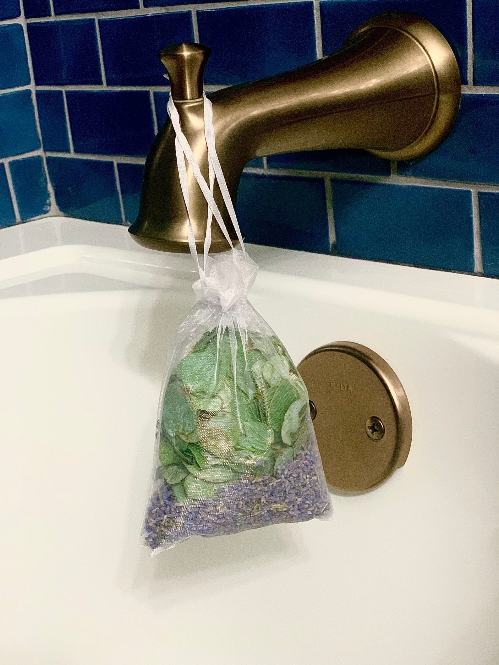 a sheer satchel with eucalyptus leaves and lavender seeds in it hanging from a bathtub faucet