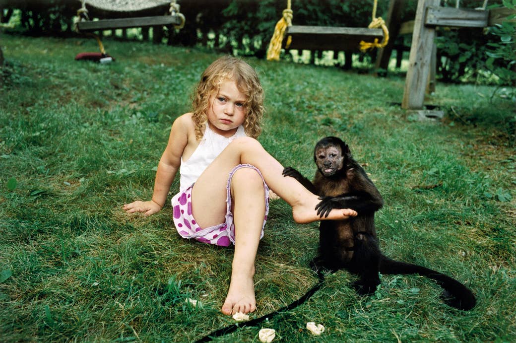 A young girl sits on the ground beside a small monkey, whose hands are on her shin and foot