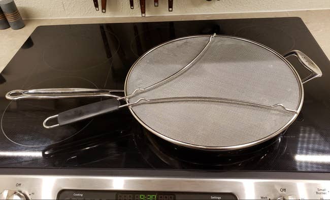 A reviewer photo of a stainless steel splatter guard sitting on top of a pan on a stove
