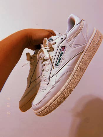 a reviewer holding up   the disconnected  achromatic  sneakers with a greenish  reebok label