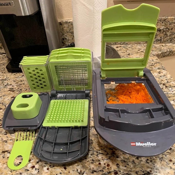 the slicer unit filled with chopped carrots with the lid opening, sitting next to all of the various blade attachments 