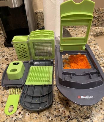 the slicer unit filled with chopped carrots with the lid opening, sitting next to all of the various blade attachments 