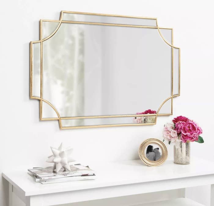 The gold mirror has overlapping detailing and is hung in a white, sunlit room
