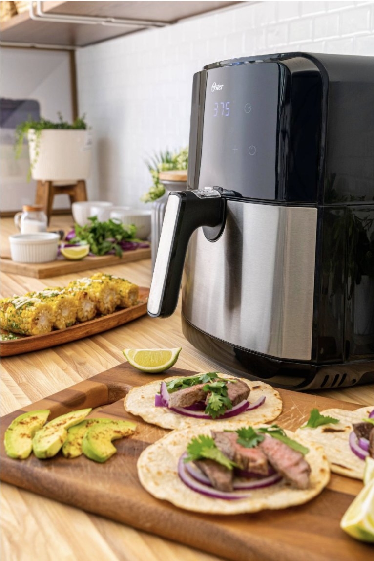 The air fryer is black with a chrome basket and handle and it&#x27;s placed atop a counter around tacos, avocado and corn 