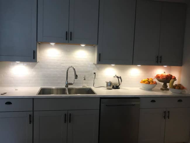 reviewer photo of a gray kitchen with multiple puck lights lighting up the counter underneath the cabinets