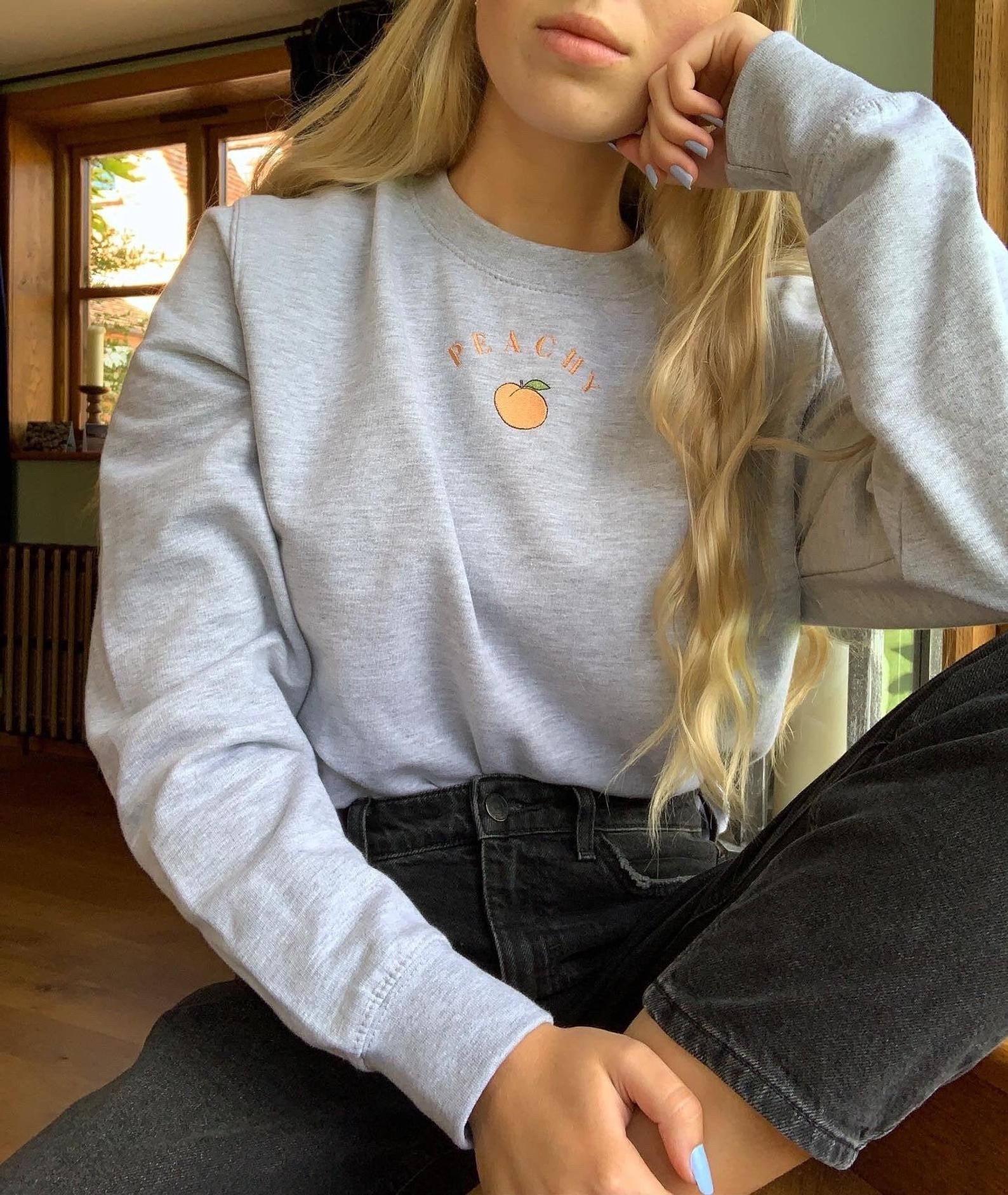 model wearing gray crew neck sweater with tiny peach embroidered on front with words &quot;peachy&quot;