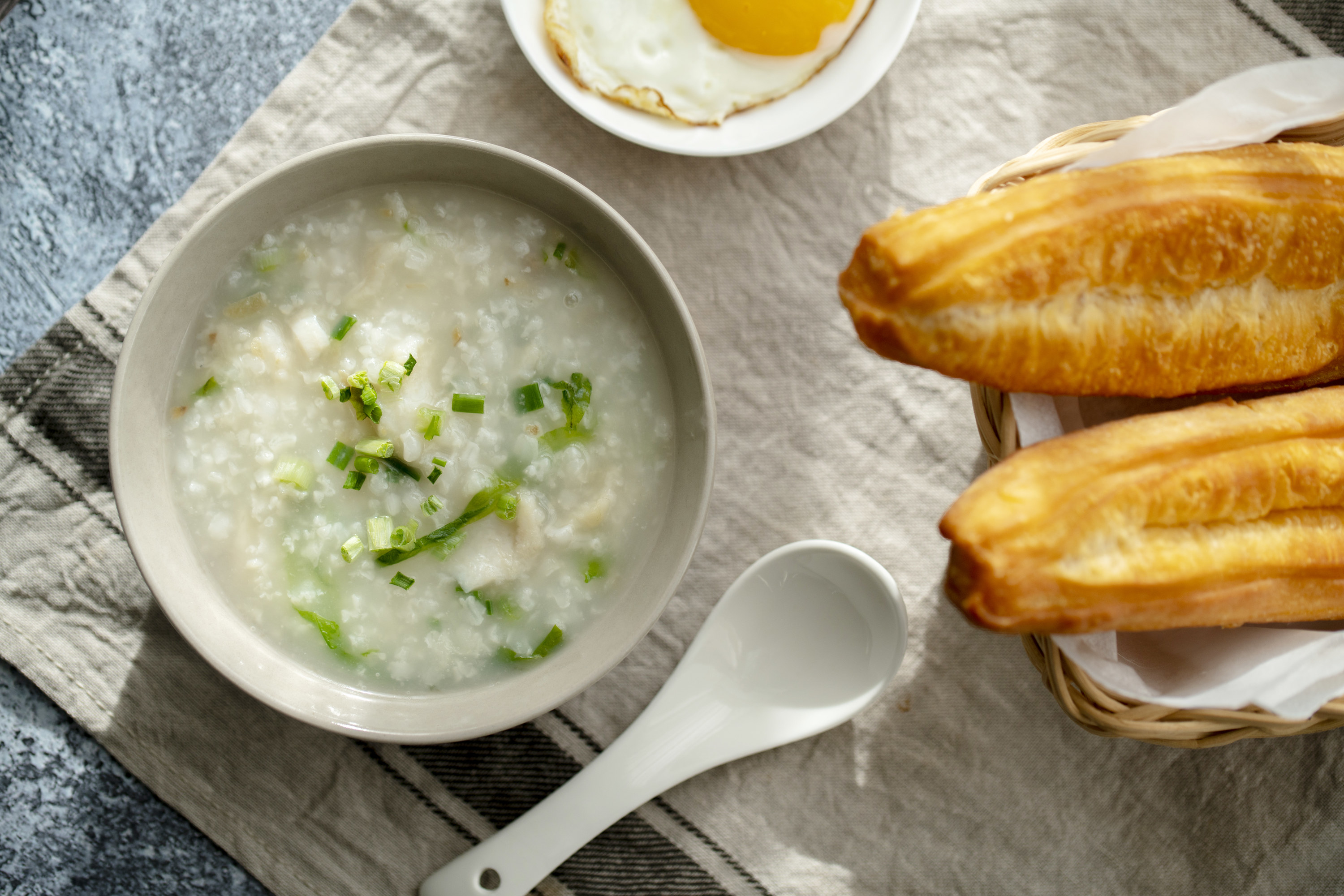 Congee in a bowl with a white soup spoon nearby with eggs and bread off to the side