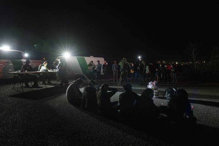 Border Patrol agents sit at a table in front of their vehicles while asylum-seekers sit on the pavement waiting to be processed