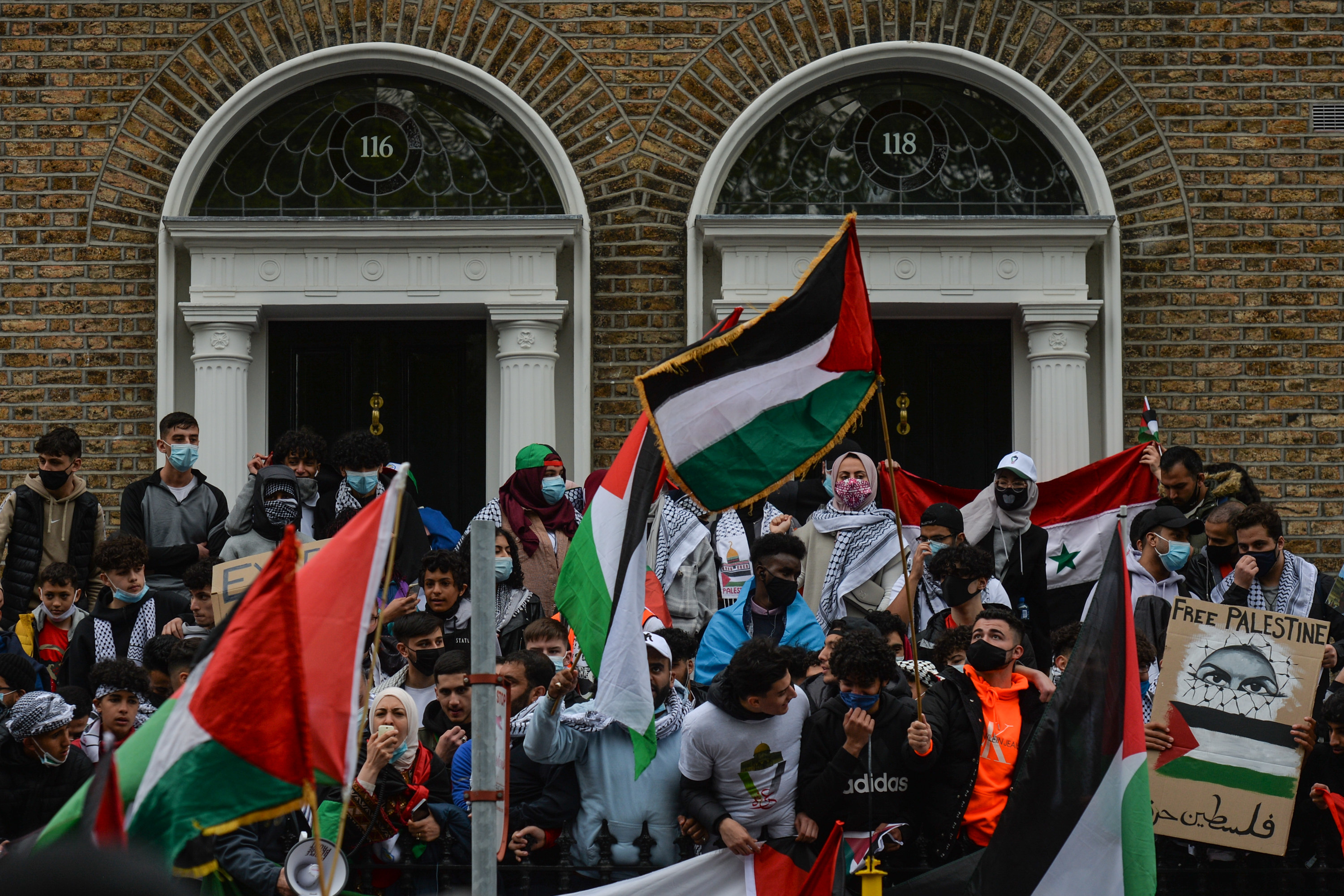 Protesters, many wearing masks, hold up Palestinian and other flags and a &quot;Free Palestine&quot; sign