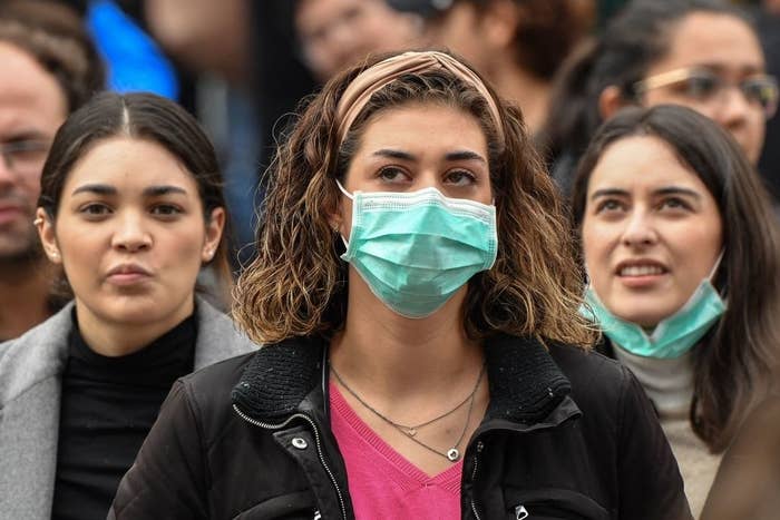 Three women in a crowd, one not wearing a mask, one wearing a surgical mask, and one wearing a surgical mask below her chin