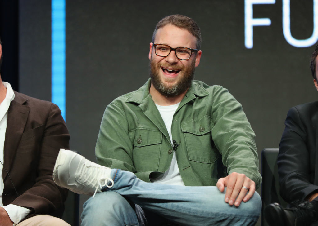 Seth, dressed casually, sitting on a panel, and laughing, with one foot resting on the opposite knee