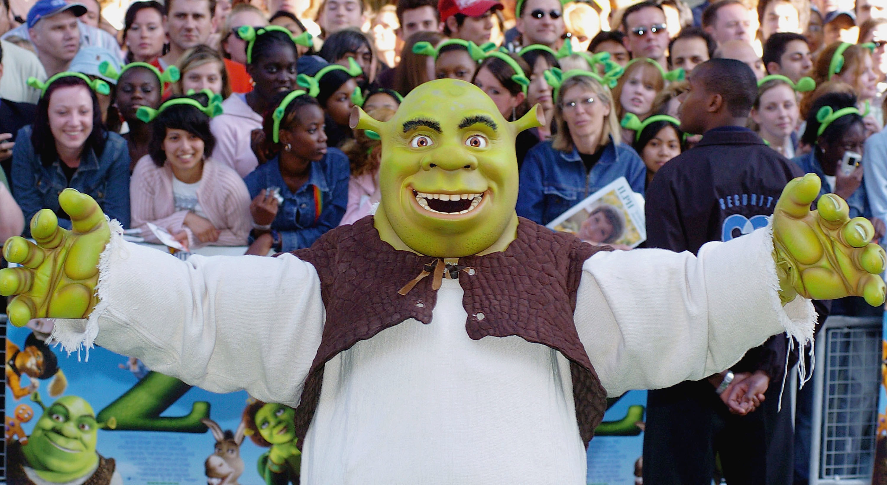 Someone in a Shrek costume holds their arms wide in front of a crowd of people wearing green headbands of ogre ears