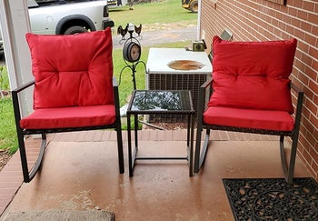 a reviewer photo of a pair of rocking chairs with red cushions with the matching side table between 