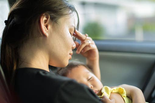 A woman holding a baby has her hand to her head. She appears tired and worried.