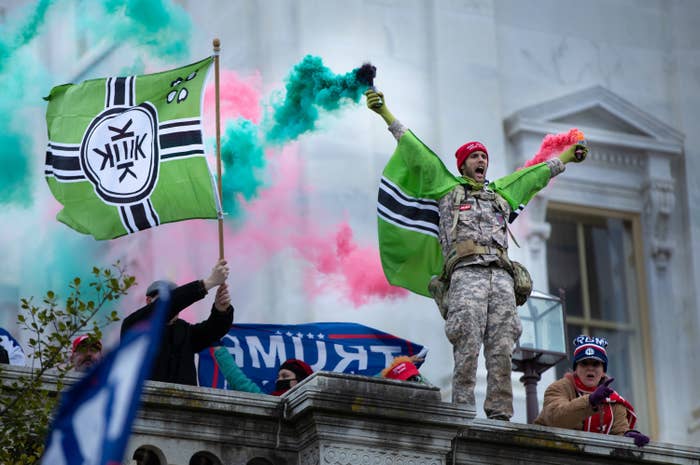 A man dressed in a military-style uniform with a flag draped over his shoulders stands on a ledge and holds a colored smoke canister in each hand