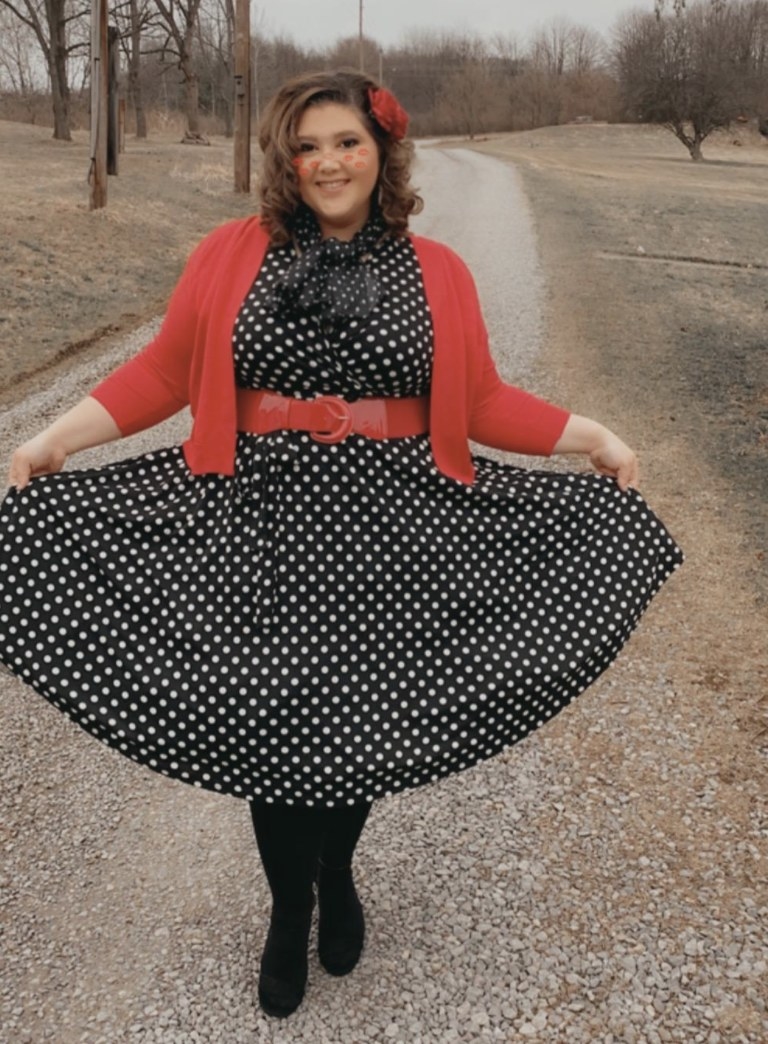 A reviewer wearing a black dress with white dots and a red cardigan