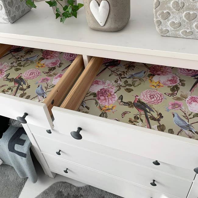 A partially open drawer revealing a floral and bird design liner inside a white dresser with silver knobs