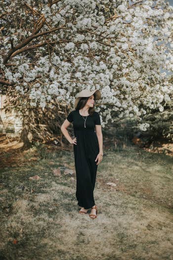 a reviewer standing in front of a spring floral tree in a black maxi dress