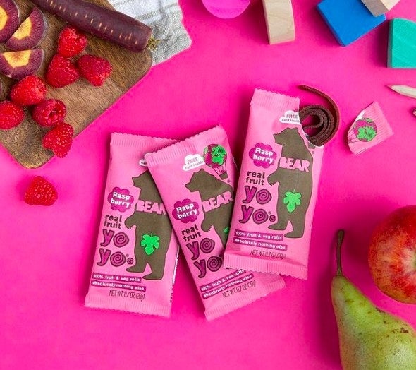 Raspberry BEAR fruit rolls are in a pink package with a cartoon bear illustration and the words &quot;real fruit yo yo&quot;. One pack is open to reveal one roll-up strip. In the background, there are real raspberries, purple carrots, an apple, and pear