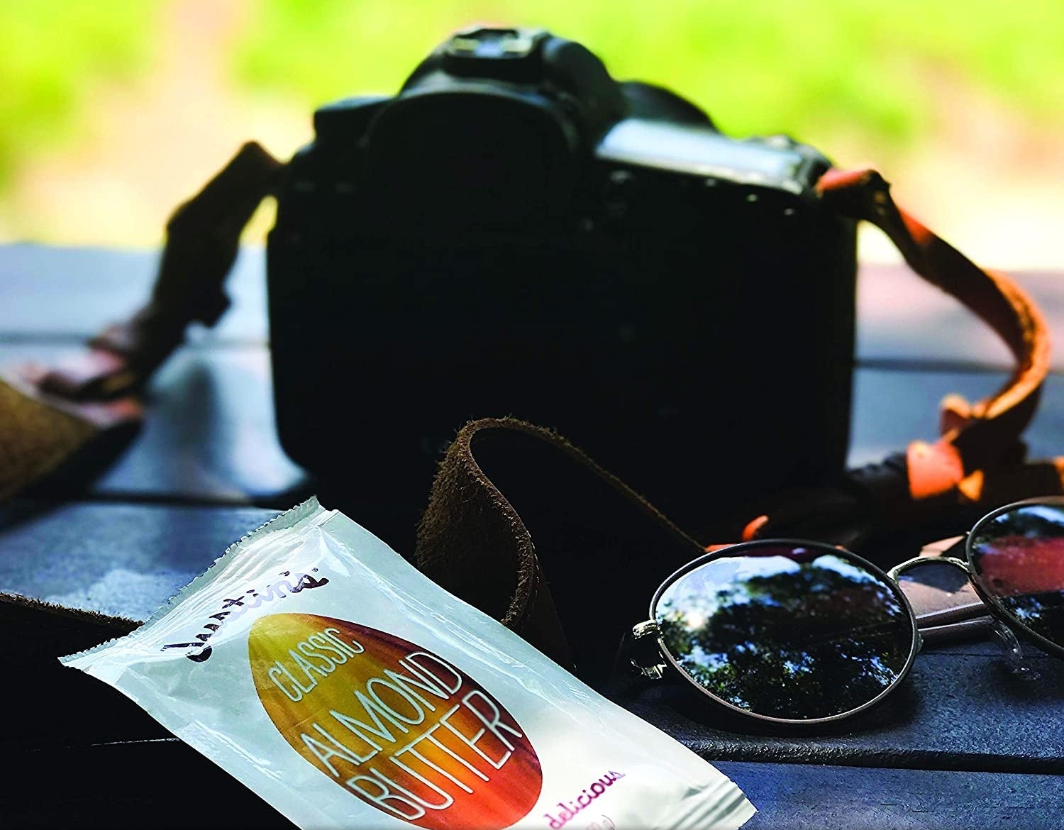 Packet of almond butter propped against a digital camera and next to a pair of sunglasses. All three objects rest on a blue picnic table. Product infographics display 6g protein, Orangutan friendly palm oil, Non-GMO verified, GF, and U