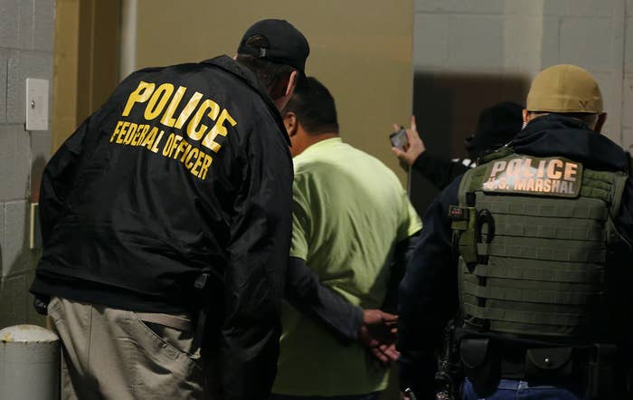 Two officers wearing clothing labeled "police US marshal" and "police federal officer" escort someone with their hands handcuffed behind their back