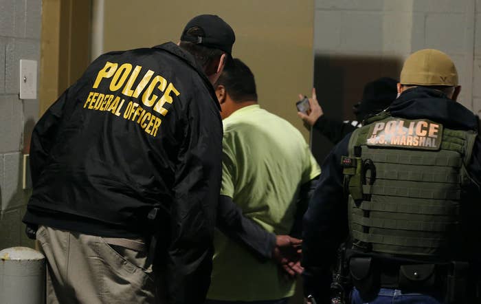 Two officers wearing clothing labeled &quot;police US marshal&quot; and &quot;police federal officer&quot; escort someone with their hands handcuffed behind their back