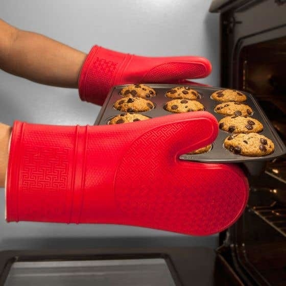 A person wearing oven mitts and taking a tray of muffins out of the oven