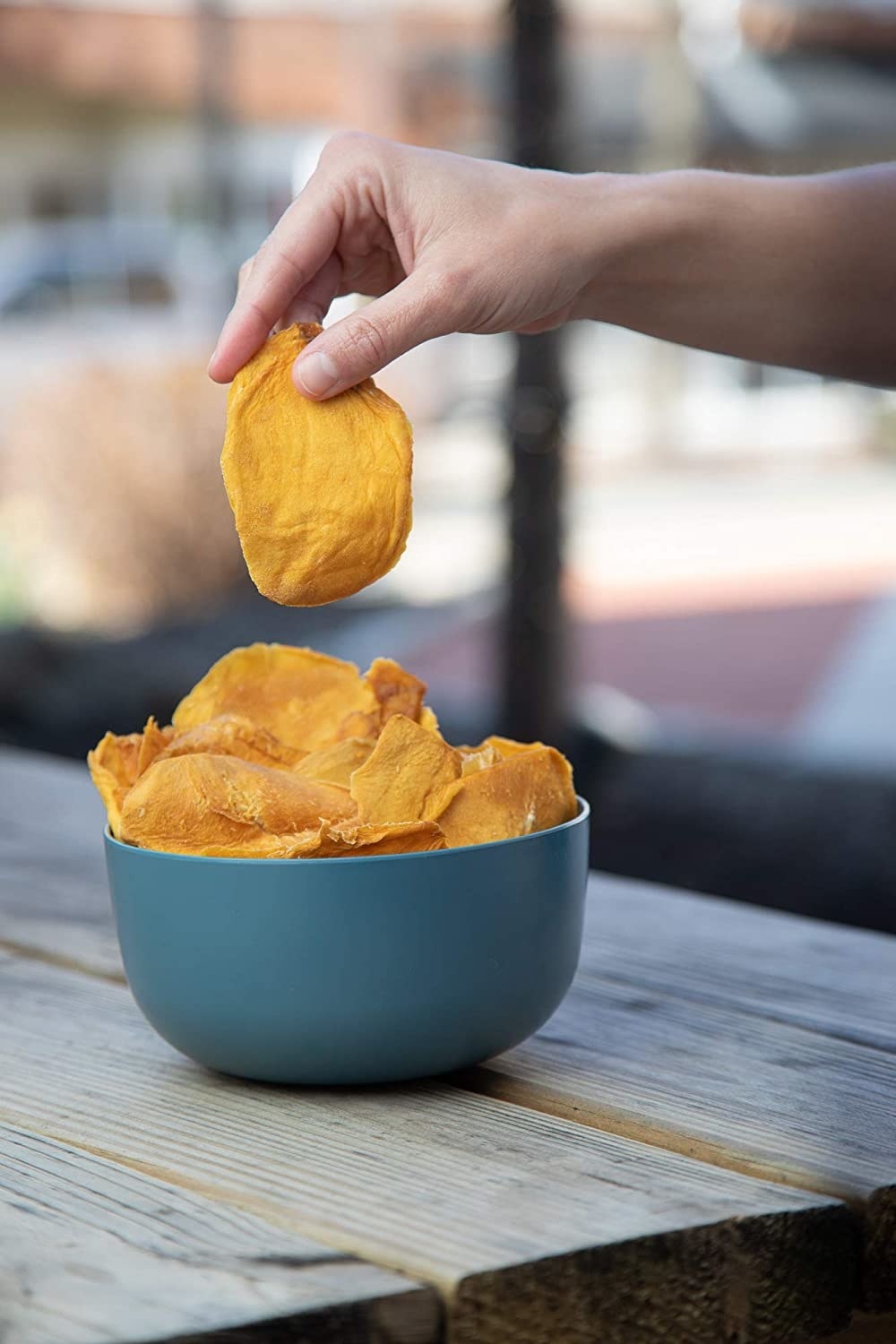 The dried mango in a blue bowl, with someone&#x27;s hand holding a single piece above the bowl to portray it&#x27;s size and texture