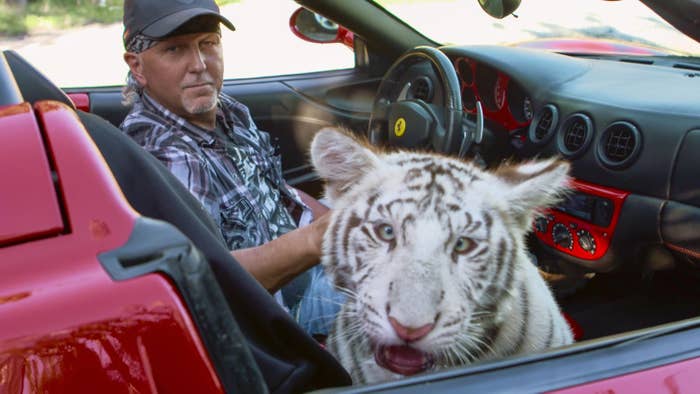 A man in the driver&#x27;s seat of a sports car and a white tiger in the passenger seat look at the camera