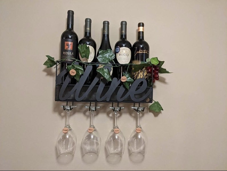 A metal wine container that reads &quot;wine&quot; mounted on a wall with clear wine glasses and faux ivy 