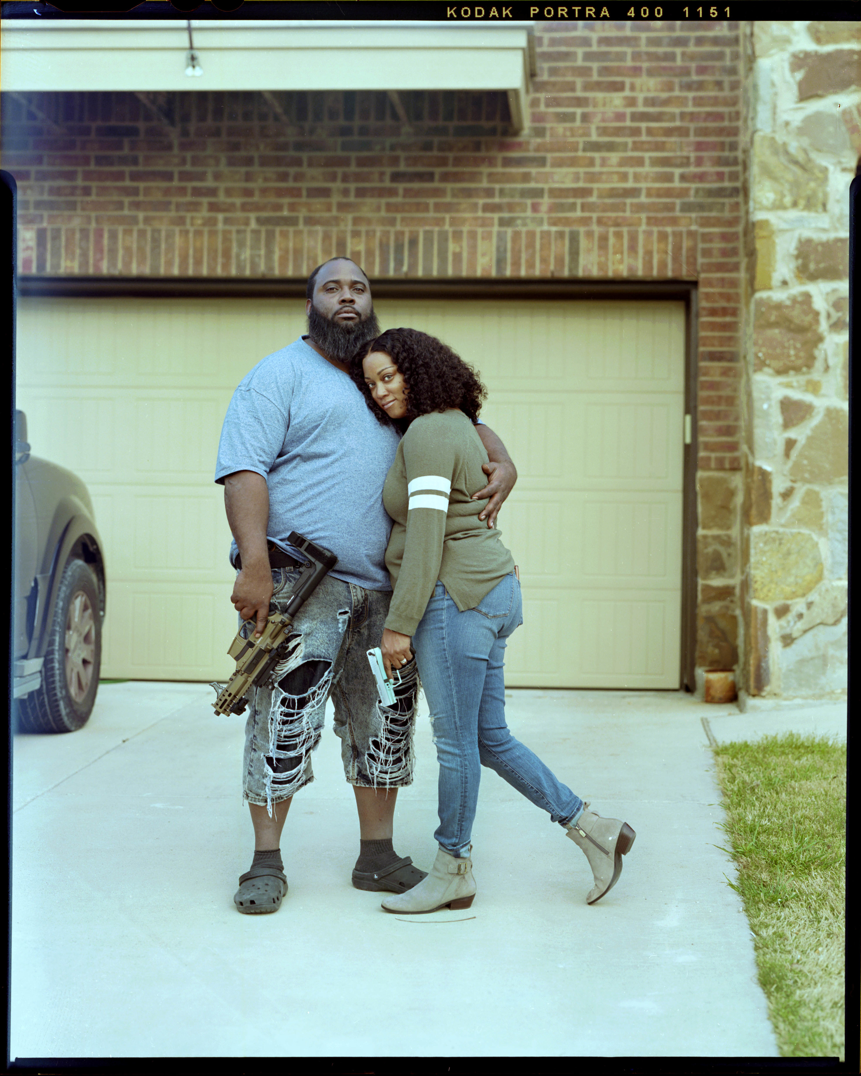 A couple embracing in front of their garage, each is holding a gun which they display for the camera